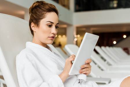 attractive young woman in bathrobe using digital tablet while relaxing in spaの写真素材