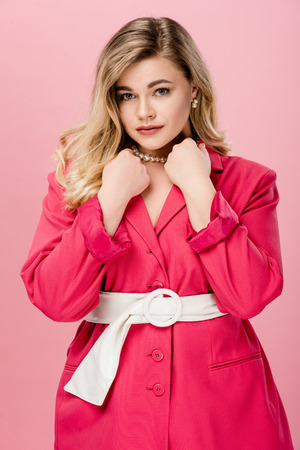 elegant young woman in pink trench coat looking at camera isolated on pinkの写真素材