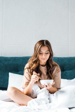 beautiful girl holding bowl with breakfast while sitting on bedの写真素材