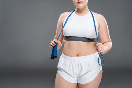 cropped shot of young oversize woman in sportswear holding jumping rope on grey