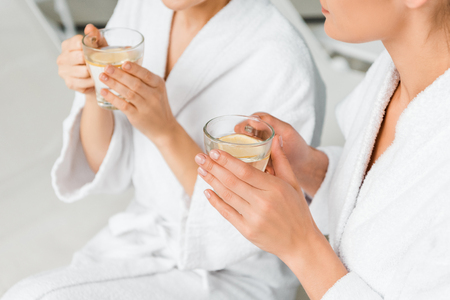 Cropped shot of young women in bathrobes holding cups with herbal tea and lemon while resting in spaの写真素材