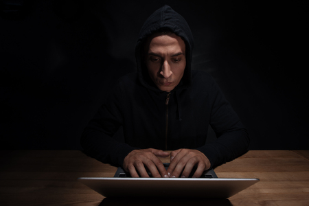 Portrait of concentrated hacker in black hoodie using laptop, cuber security conceptの写真素材