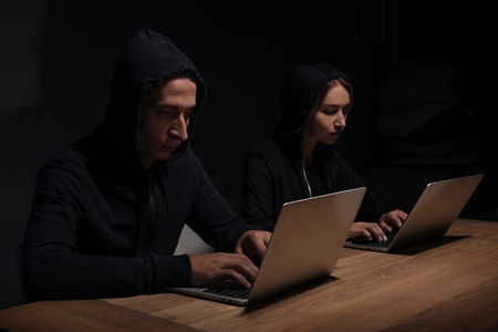 Focused hackers in black hoodies using laptops in dark room, cyber security conceptの写真素材