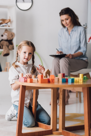 Happy little child playing with blocks and looking at camera while psychologist sitting blurred on backgroundの写真素材