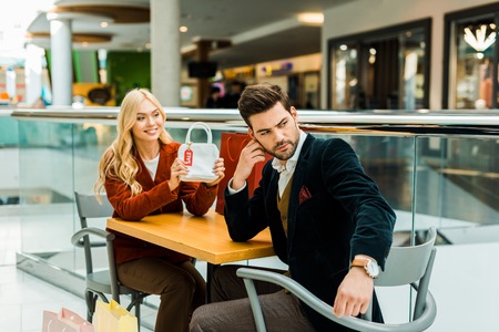 Female shopaholic showing bag with sale tag to upset boyfriend in shopping center