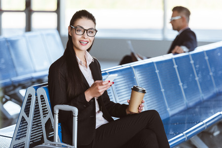 Young smiling businesswoman in glasses with coffee to go using smartphone in departure lounge at airportの写真素材