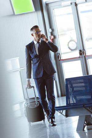 Adult handsome businessman with smartphone and baggage walking at airportの写真素材