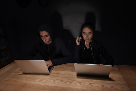 Hackers in black hoodies using laptops at wooden tabletop in dark room, cyber security conceptの写真素材