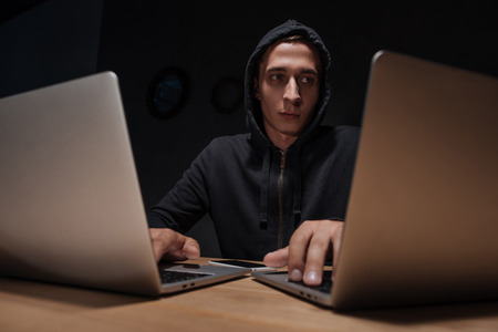 Portrait of hacker in black hoodie using laptops in dark room, cyber security concept
