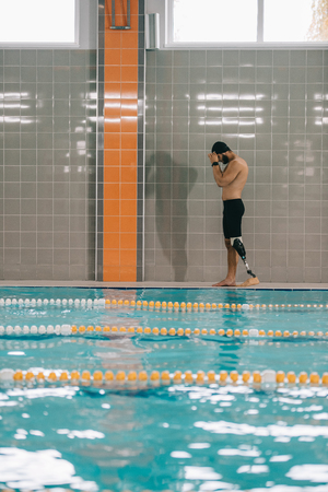 Handsome young sportsman with artificial leg walking by poolside at indoor swimming poolの写真素材