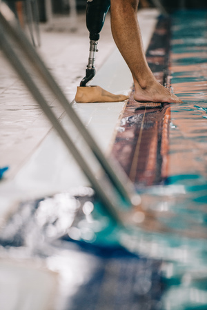 Cropped shot of swimmer with artificial leg standing on poolside at indoor swimming pool and checking water temperatureの写真素材