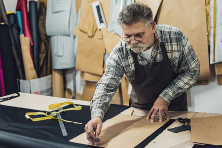 Serious mature male leather handbag craftsman in apron and eyeglasses working at studioの写真素材