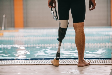 Cropped shot of swimmer with artificial leg standing in front of indoor swimming pool and holding swimming gogglesの写真素材