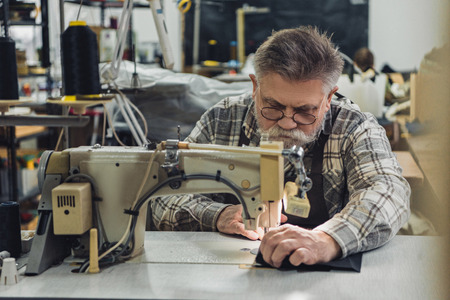 Focused mature male tailor in apron and eyeglasses working on sewing machine at studio
