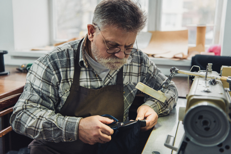 Mature male tailor in eyeglasses and apron cutting leather by scissors at workshop