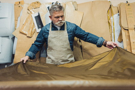 Selective focus of middle aged male handbag craftsman putting fabric on working table at studioの写真素材