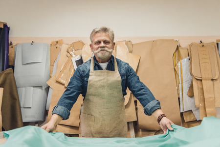 Cheerful mature male handbag craftsman putting fabric on working table at studio