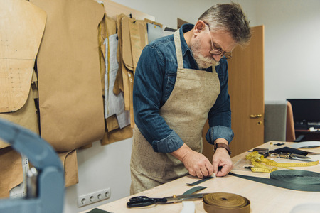 Selective focus of middle aged male handbag craftsman in apron and eyeglasses working with fabric at studioの写真素材