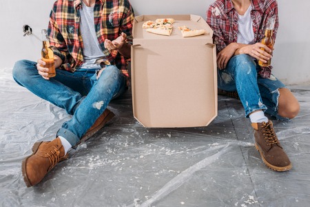 Cropped shot of young couple holding beer bottles and sitting with pizza box in new apartment