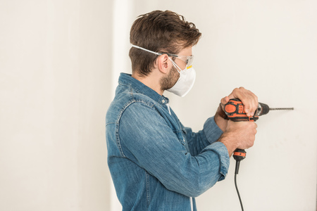 Young man in protective work wear using electric drill at wall during house repairの写真素材