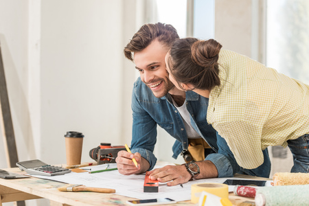 Young woman kissing happy boyfriend marking blueprint with level tool during renovationの写真素材