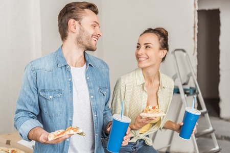 Happy young couple holding paper cups with drinking straws and pizza slices in new apartmentの写真素材