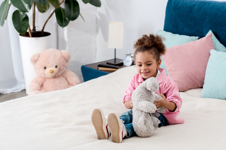Smiling African american child sitting on the bed and playing with her teddy bearの写真素材