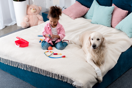 Cute African american child playing with toys on the bed with white dog lying besideの写真素材