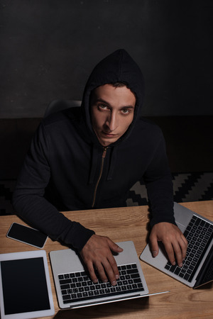 High angle view of male hacker in black hoodie using laptops at tabletop with tablet and smartphone, cyber security conceptの写真素材