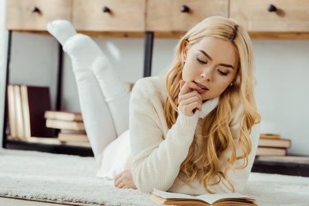 Thoughtful attractive woman laying on carpet and reading bookの写真素材