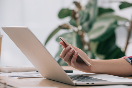 Cropped shot of businesswoman with smartphone at workplace with laptopの写真素材