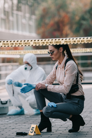 Female detective in sunglasses wearing latex gloves with criminologist behind at crime sceneの写真素材