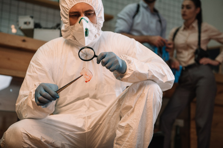 Close up of forensic investigator examining evidence with magnifying glass at crime scene with colleagues working behindの写真素材