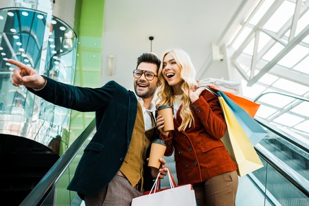 Fashionable boyfriend with coffee to go showing something to excited girlfriend with shopping bags standing on escalatorの写真素材