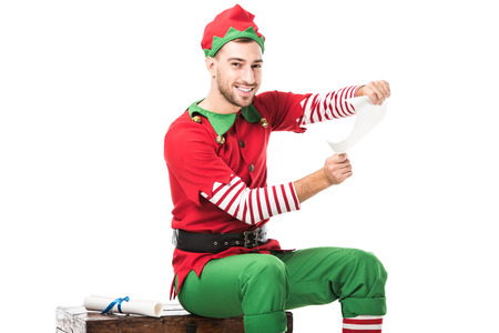 smiling man in christmas elf costume sitting and holding wishlist roll isolated on whiteの写真素材