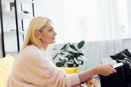 beautiful smiling mature woman sitting on bright yellow sofa and pushing buttons on tv remote controllerの写真素材