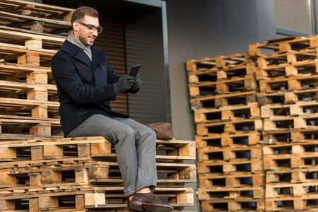 handsome smiling man using smartphone and sitting on wooden pallets
