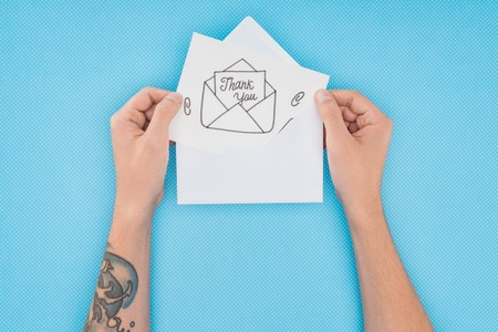 cropped person holding paper with thank you lettering sticking out of envelope isolated on blue...
