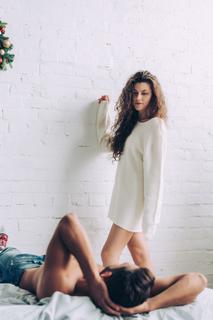 beautiful curly young woman standing near boyfriend laying on bed at homeの写真素材