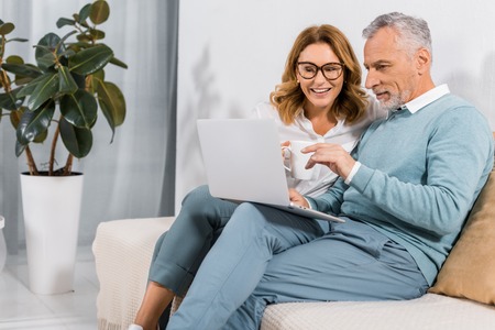 handsome mature man pointing by hand at laptop screen to beautiful wife on sofa at homeの写真素材