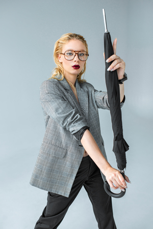 beautiful stylish girl in glasses and gray jacket posing with umbrella isolated on greyの写真素材