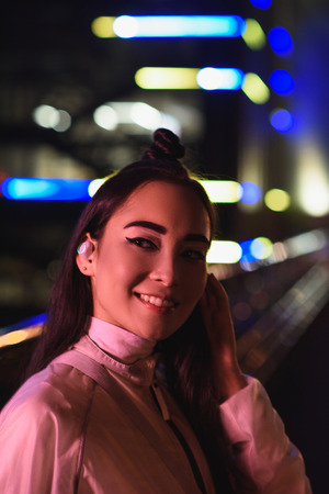 smiling stylish asian girl with wireless earphone looking away on street with neon light in evening, city of future conceptの写真素材