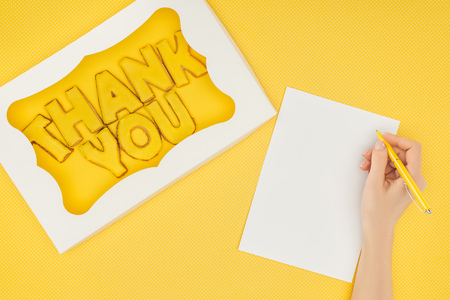 cropped person holding pen above notebook and square-shaped cake in box with thank you lettering isolated on yellow backgroundの写真素材