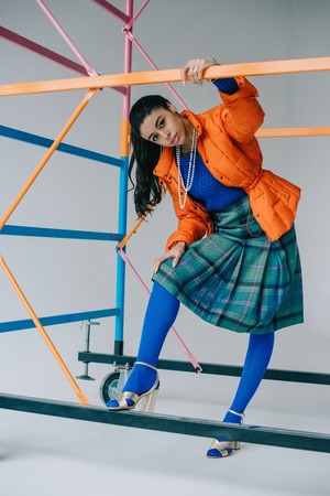 stylish girl in orange winter jacket, checkered skirt and blue tights posing near colorful scaffold in studio