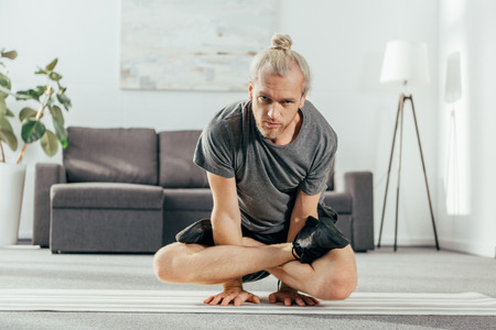adult man balancing on arms with crossed legs and looking at camera while training at homeの写真素材