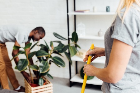 blonde woman holding mop while african american man putting plant in wooden pot