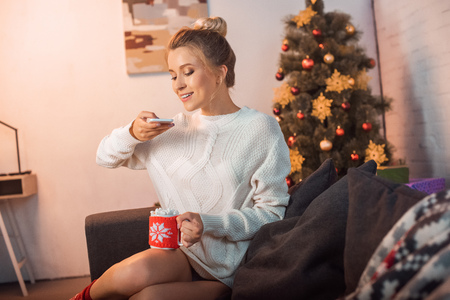 happy young blonde woman sitting on couch and taking photo of hot cocoa with marshmallows on smartphone at christmas timeの写真素材