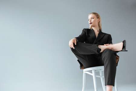 attractive fashionable girl in black suit sitting on chair isolated on greyの写真素材
