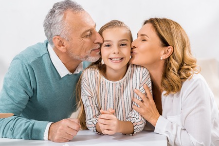 adorable child looking at camera while grandparents kissing her in cheeksの写真素材
