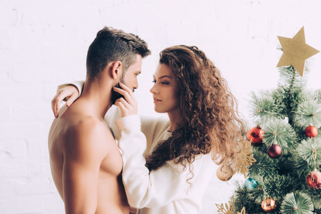 attractive young woman embracing shirtless boyfriend near christmas tree at homeの写真素材